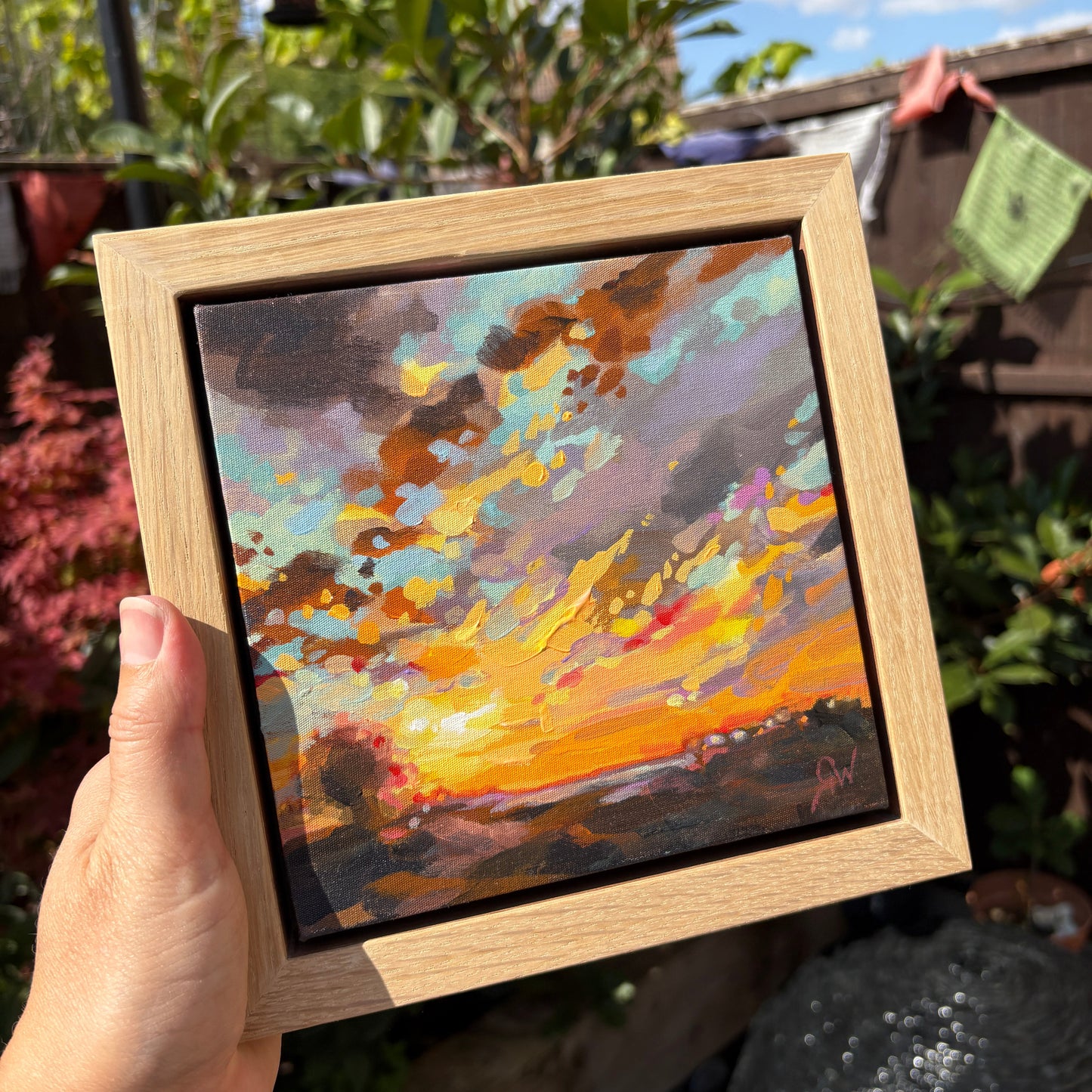 A hand holds a square framed oil painting of a vibrant sunset landscape. The painting features a bright orange and yellow sunburst, dramatic clouds in shades of brown, gray, and blue, and a dark horizon.