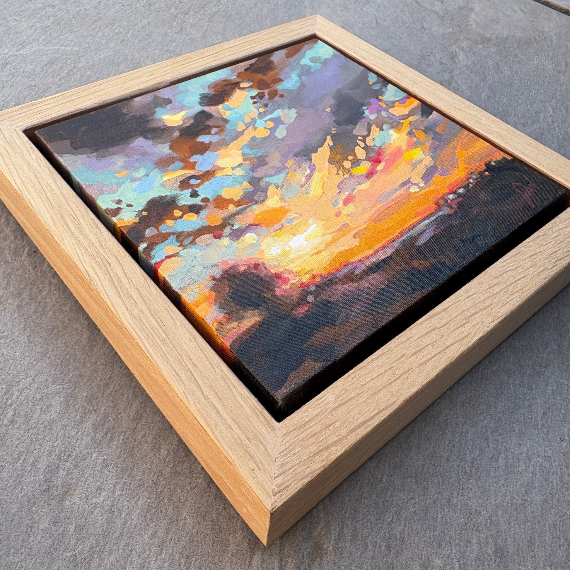 A square framed abstract landscape painting with vibrant orange, yellow, blue, and purple brushstrokes, set in a light natural wood frame on a gray stone surface.